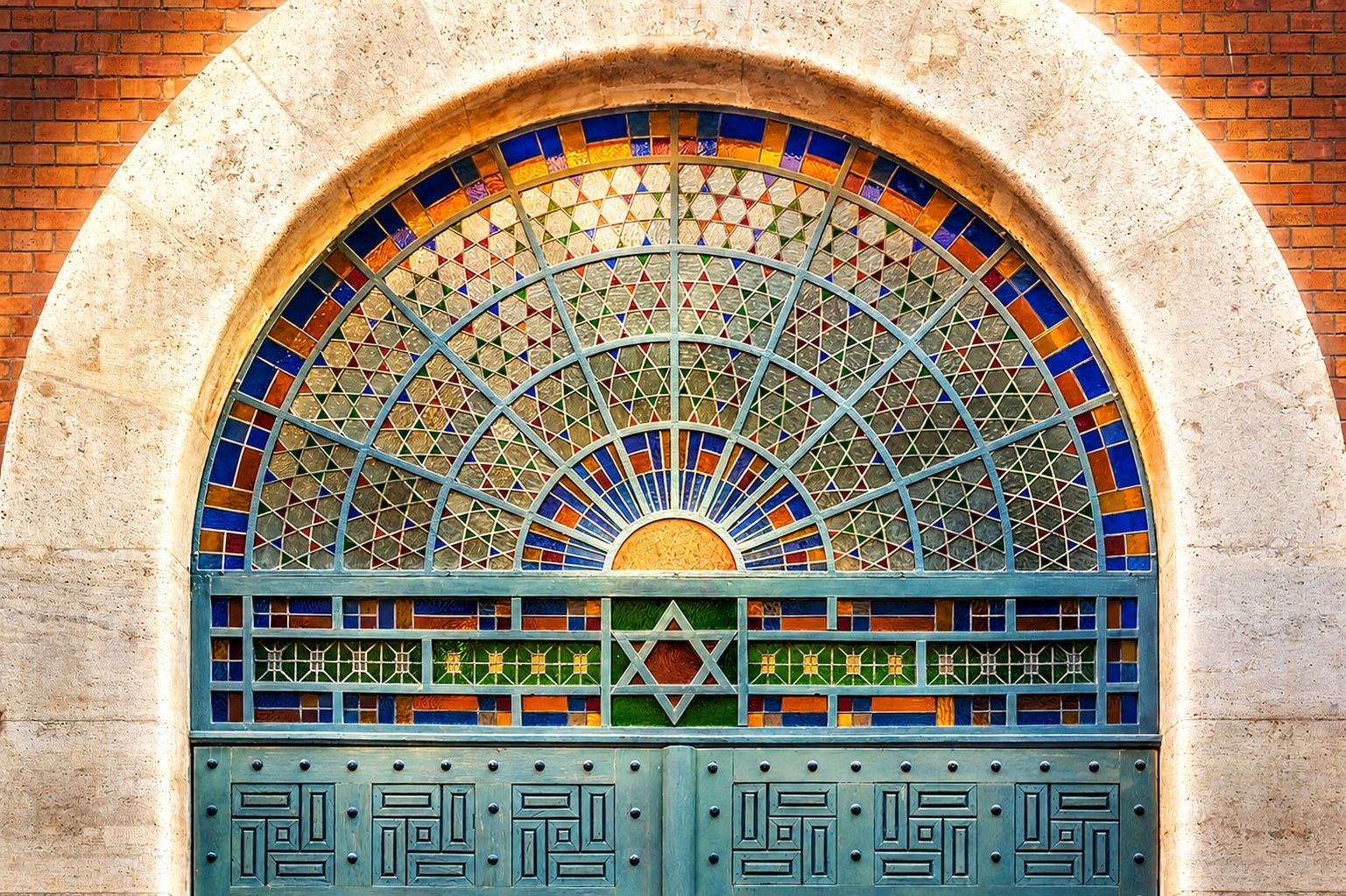 Stained glass arched window with Star of David above the painted blue doors of a Budapest synagogue, set into a brick façade.