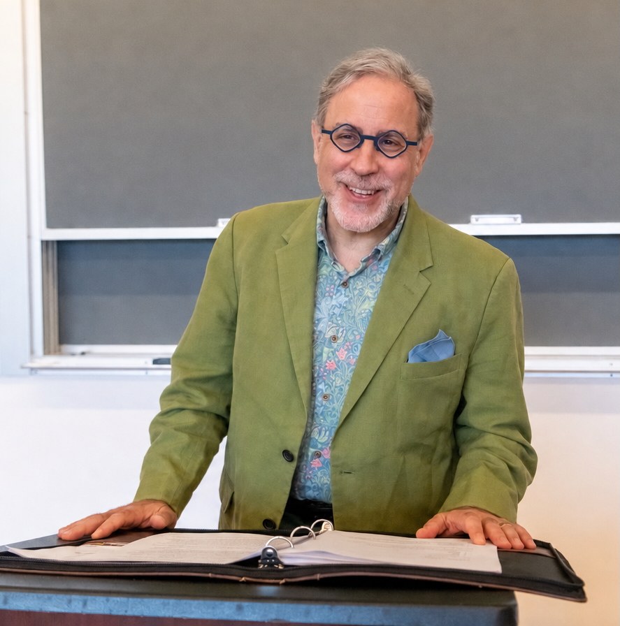 Marc Michael Epstein in a green linen blazer and patterned shirt, standing at a classroom lectern with an open binder, a chalkboard behind him.