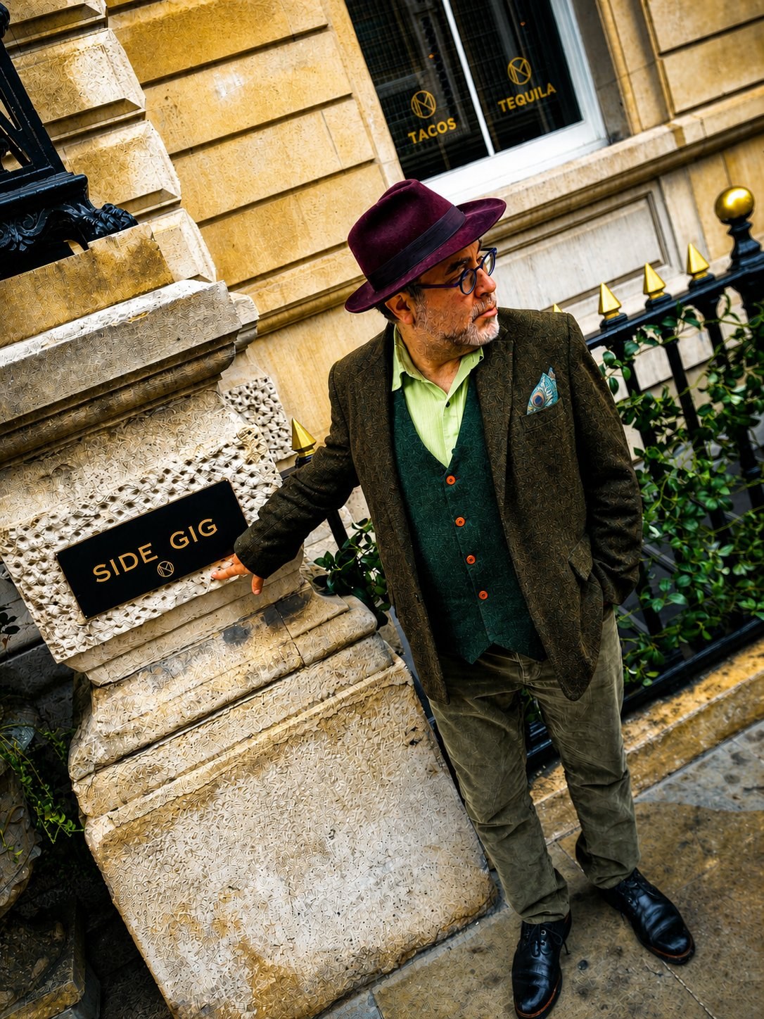 Marc Michael Epstein in burgundy fedora, tweed jacket and green corduroys, leaning against a stone plinth in front of the &lsquo;Side Gig&rsquo; restaurant, his MM&Epsilon; monogram visible on the awning above.