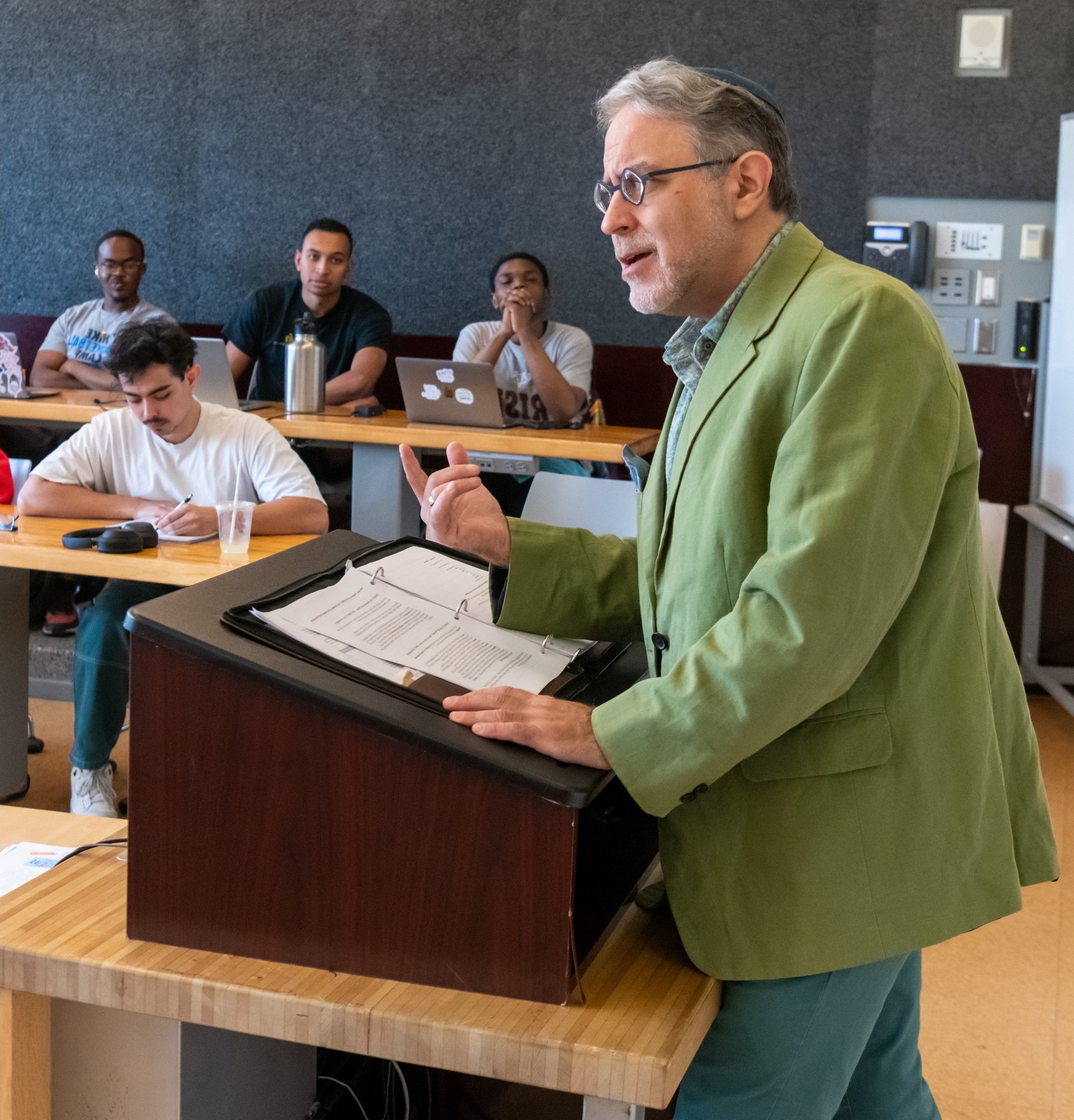 Marc Michael Epstein lecturing in a Vassar classroom, gesturing from a wooden lectern with an open binder, students at tables behind him.